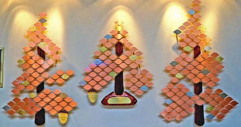 The Giving Tree: three wall-mounted tree sculptures made of copper and colored leaf plaques with brass pine cones, lit by spotlights on a white wall.